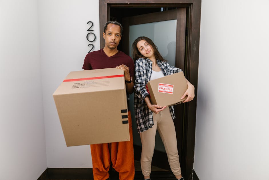 Two individuals, a man wearing a maroon t-shirt and orange trousers and a woman dressed in a plaid shirt and beige pants, stand inside a narrow corridor near a doorway with a dark wooden frame. They are holding several cardboard boxes, one with visible red and white 'Fragile' label and another with blue markings, indicating their use during packing and moving. The man is gripping a large, rectangular box with a red strip across the top, while the woman cradles a smaller box close to her chest. Behind them, the door is slightly ajar, revealing a glimpse of the room beyond. The setting suggests a home relocation process, with the individuals preparing to navigate a narrow doorway or staircase, typical of flats needing careful furniture transport. The environment appears well-lit, with clean white walls and a simple interior that highlights the moving activity, reflecting the logistics involved in house removals and packing for a move, as managed by Islington Removals.