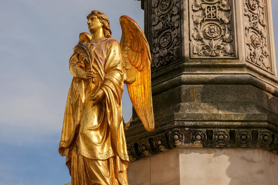 Close-up of a gilded statue of an angel holding a bundle of wheat, situated on the corner of an ornate stone building. The angel has large, detailed wings painted in a warm, bronze tone, and is dressed in flowing robes with intricate folds. The statue is positioned near the base of the building's decorative column, which features elaborate floral and geometric carvings. The scene is captured during daylight with a soft blue sky in the background. The setting suggests an exterior perspective, possibly part of a historic or religious structure. This type of architectural detail may be encountered during house or property relocations, as part of the delicate handling of architectural features during furniture transport or moving preparations, with Islington Removals frequently involved in such home relocation processes.