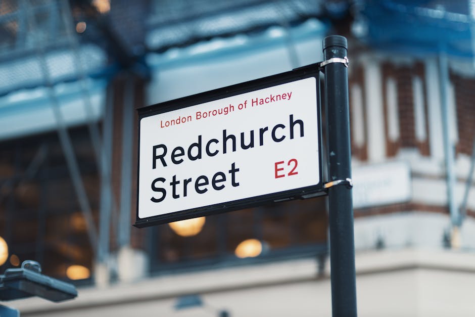 Close-up of a street name sign reading 'Redchurch Street, E2' located in the London Borough of Hackney, mounted on a black metal post with a perforated blue metal structure overhead. The sign has a white background with black text, and the street name is bold and centrally positioned. In the background, blurred urban buildings with brick facades and windows can be seen, suggesting a busy city environment. This scene captures the typical setting where house and furniture removals, such as those by Islington Removals, might be arranged, emphasizing the logistics involved in home relocation and furniture transport within London neighborhoods.